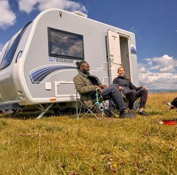 two people sitting outside a Bailey Discovery touring caravan in a green field with a cloudy blue sky