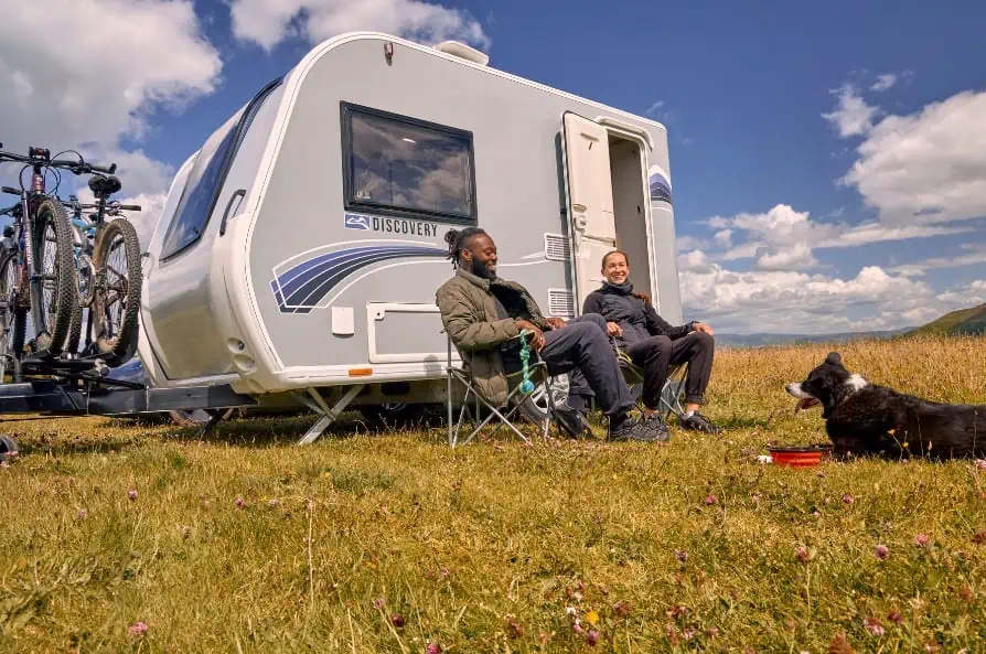 two people sitting outside a Bailey Discovery touring caravan in a green field with a cloudy blue sky